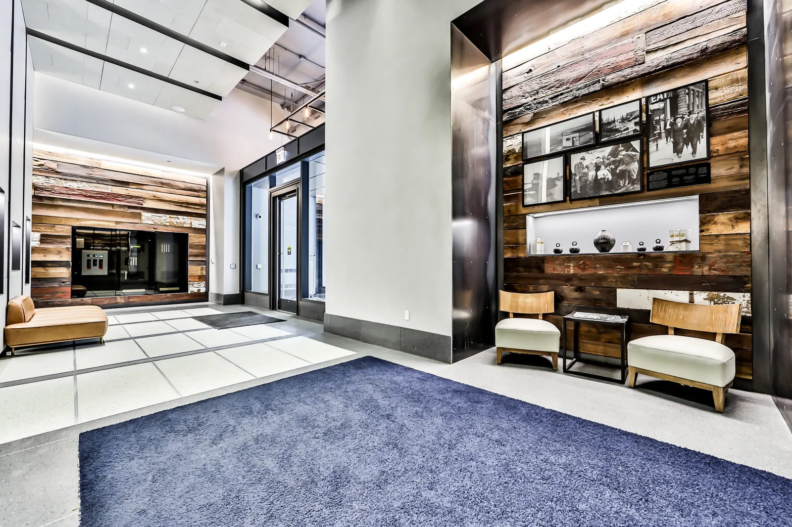 Lobby with a reclaimed-wood display wall, framed photographs, seating, and elevator doors.